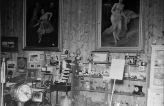 Interior view of the old Dawson City Museum located in the Firehall. Several artifacts, curios, and cupboards are visible. Two large oil paintings of nude women are hanging behind display. Items were subsequently damaged or lost in the fire.