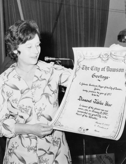 One black and white photograph of Dawson City mayor, Yolanda Burkhard, holding an official proclamation of the beginning of the City of Dawson's Diamond Jubilee Year. The proclamation is dated January 9, 1977.