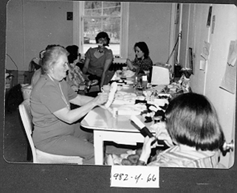 a black and white photo of six people around a table, working on the jubilee dolls. 